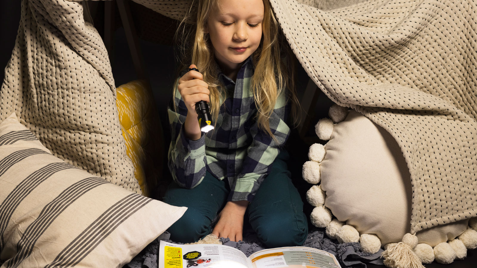 Lapsi lukee aikakauslehteä taskulampun valossa vilteistä tekemässään majassa.
