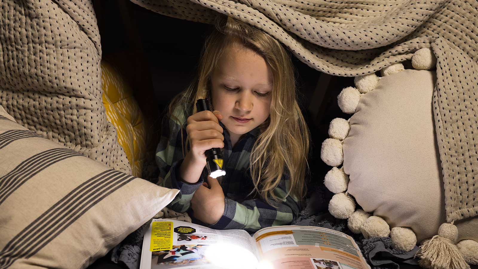 Lapsi lukee aikakauslehteä taskulampun valossa itse tehdyssä majassa.